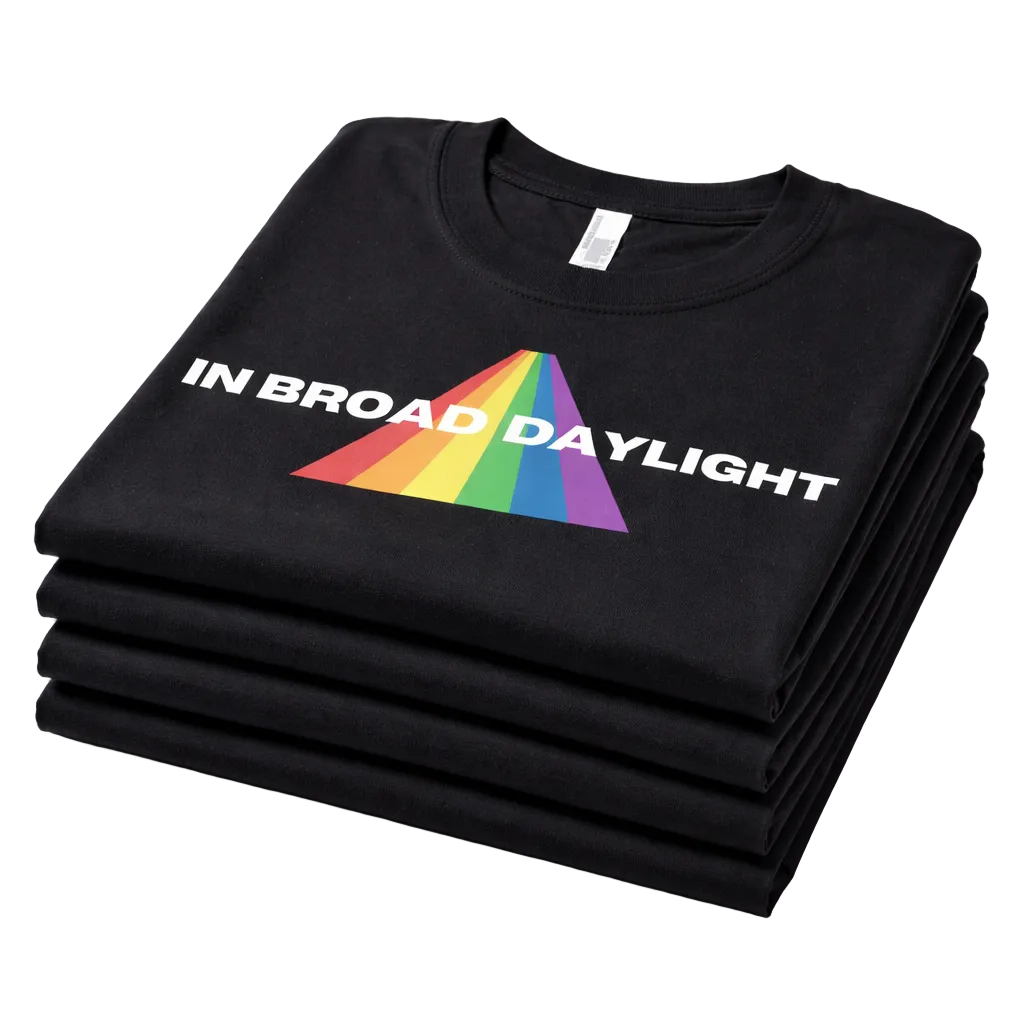 Stack of black shirts with a rainbow triangle logo and 'IN BROAD DAYLIGHT' text on a white background